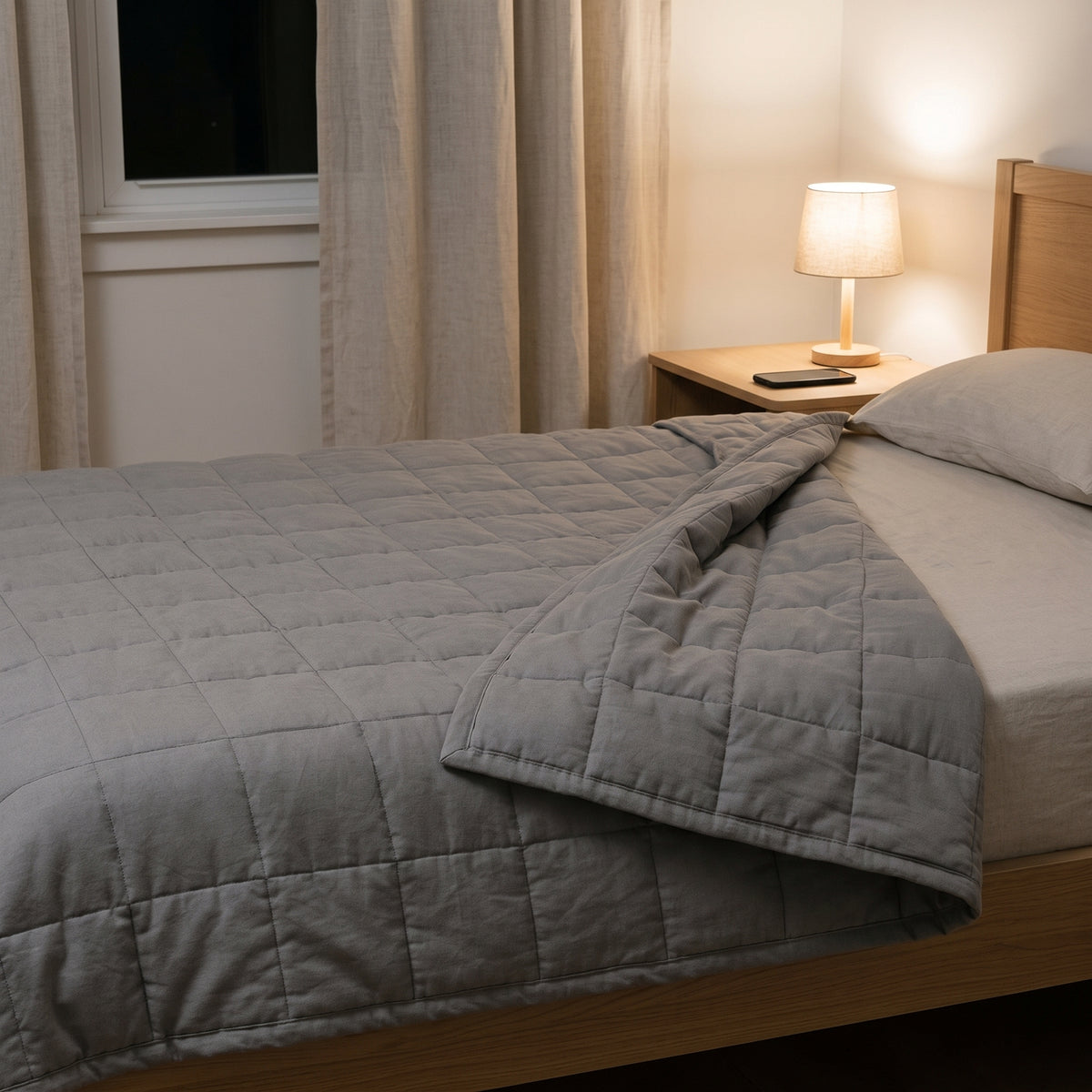 Couverture lestée Lestona grise, dépliée sur un lit dans une chambre le soir, lampe de chevet allumée